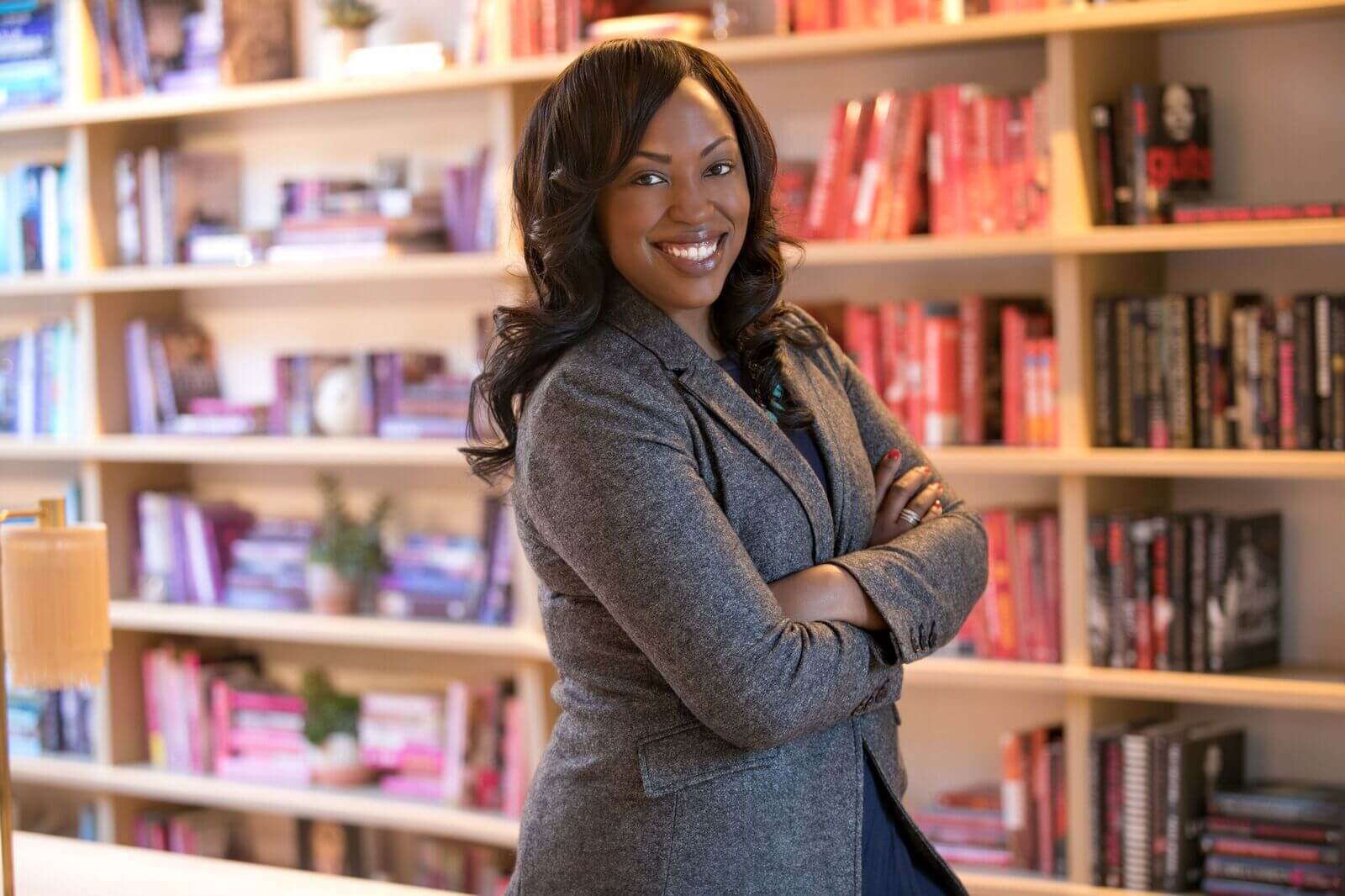 A black woman with long black hair in a gray blazer smiling with her arms crossed across her chest standing in front of a book shelf lined with red books