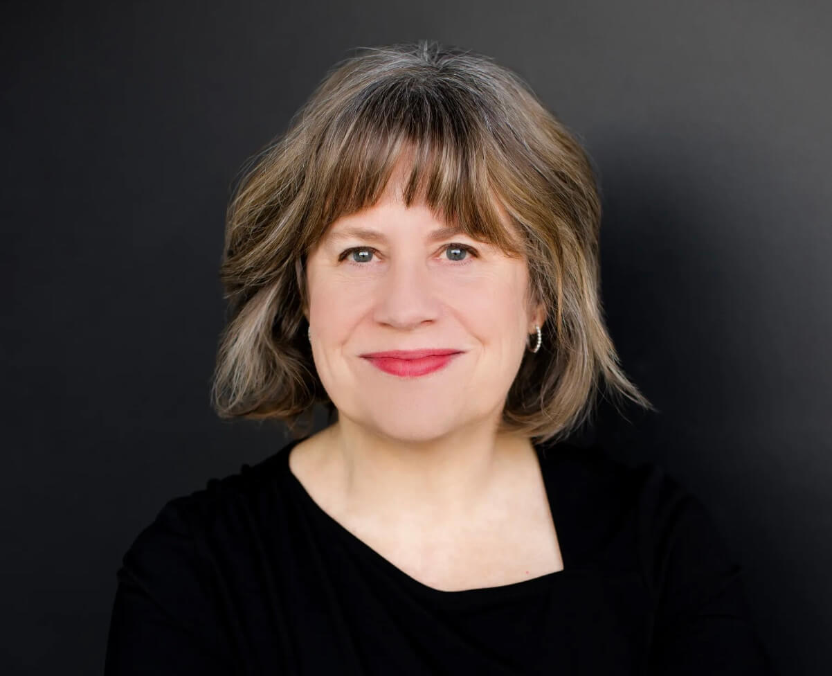 Image of a white woman with chin-length brown and gray hair wearing a black shirt and red lipstick on a gray background