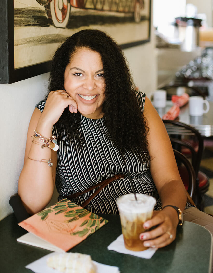A black woman with long black hair seated and leaning against a wall in a coffee shop, wearing a black and white striped sleeveless dress, holding a coffee, and smiling at the camera