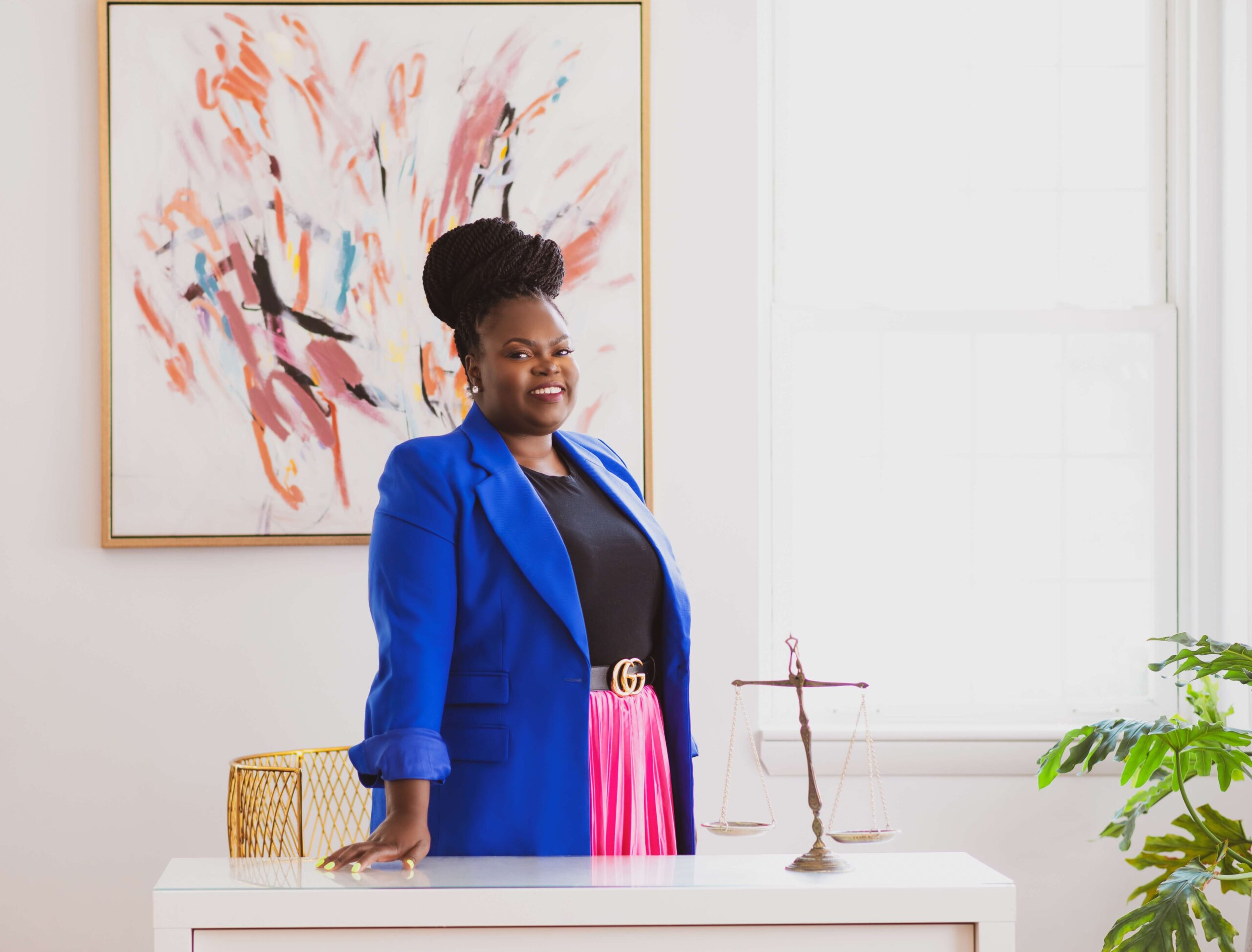 A Black woman wearing a cobalt blue blazer, black shirt and hot-pink skirt standing behind a white desk with the scales of justice and a modern abstract painting behind her smiling at the camera