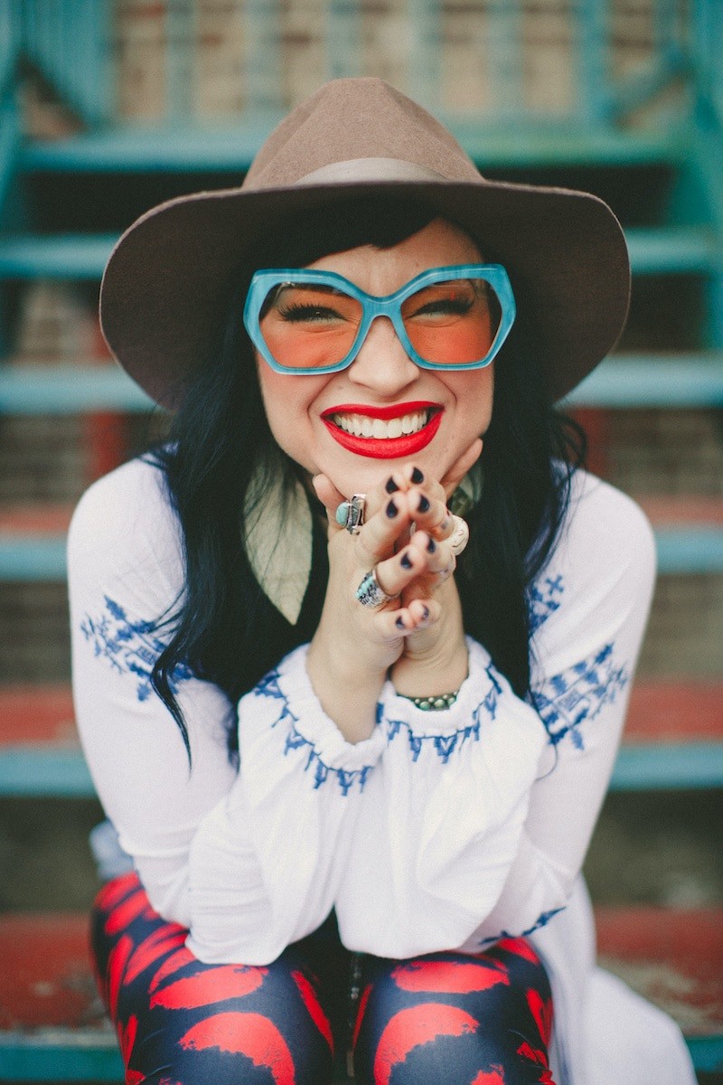 Woman in oversize teal sunglasses, a large brown cowboy hat and her hands on her chin with her elbows resting on her knees, in a white long-sleeve shirt and red leggings sitting on steps smiling at the camera with red lipstick